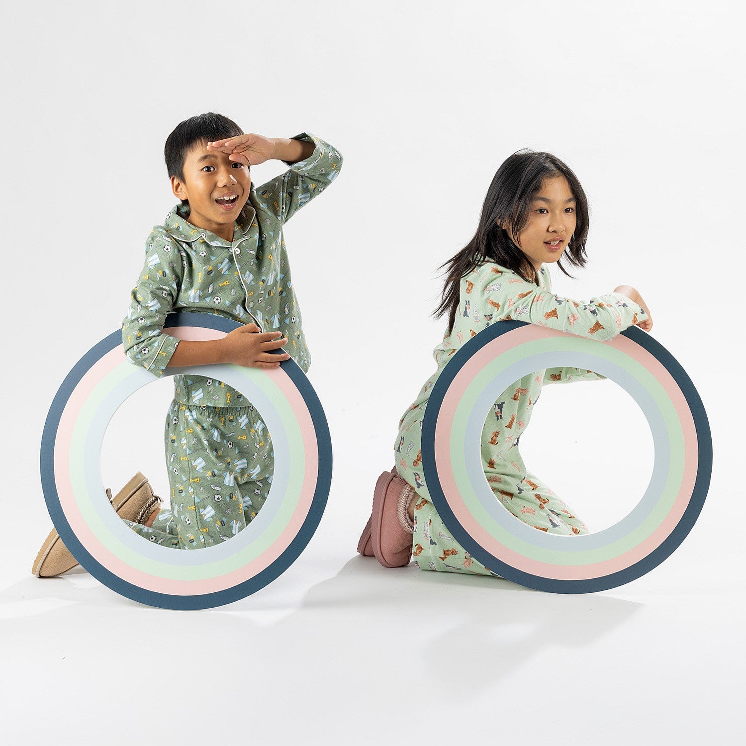 Two children wearing playful printed pyjama sets, one in green and one in cream, holding picture frame props.