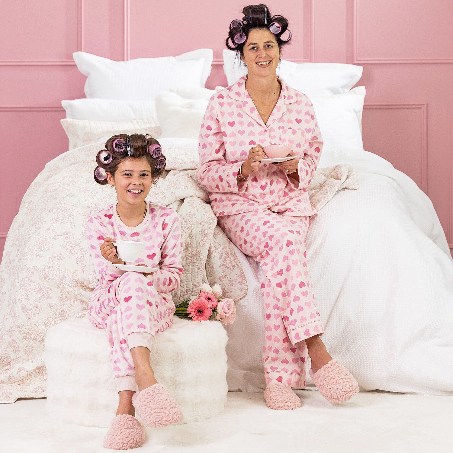 A kid, and mother wearing matching pink heart print pyjamas, sitting on a white bed with curlers in their hair.