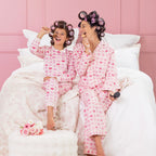 A kid, and mother wearing matching pink heart print pyjamas, sitting on a white bed with curlers in their hair.