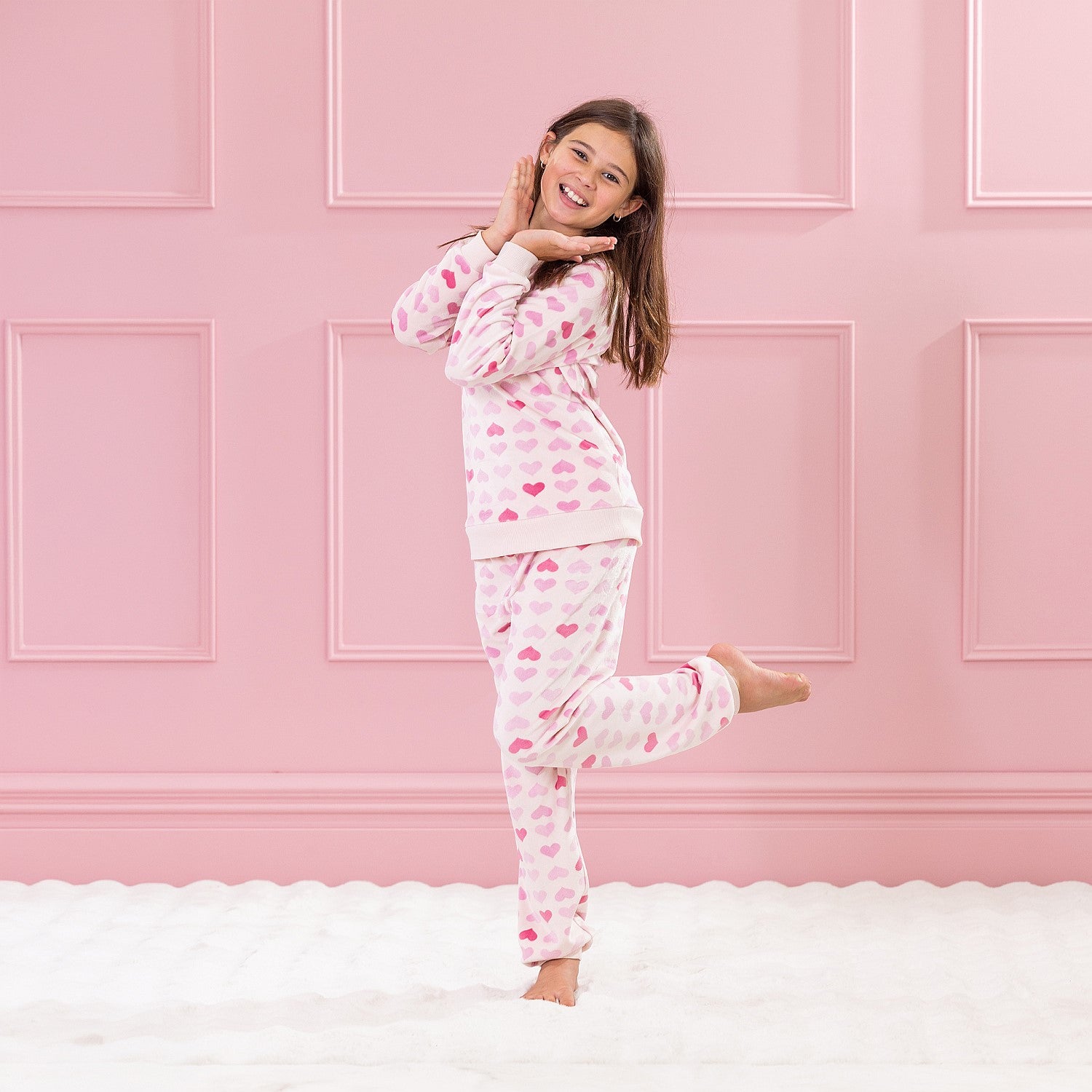 Child wearing pink heart print pyjamas standing against a pink panelled background.