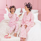 A kid, and mother wearing matching pink heart print pyjamas, sitting on a white bed with curlers in their hair.
