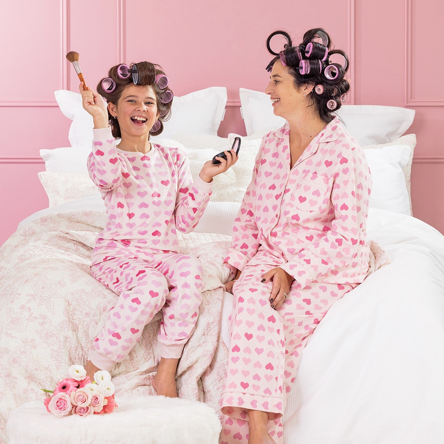 A kid, and mother wearing matching pink heart print pyjamas, sitting on a white bed with curlers in their hair.