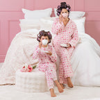 A kid, and mother wearing matching pink heart print pyjamas, sitting on a white bed with curlers in their hair.