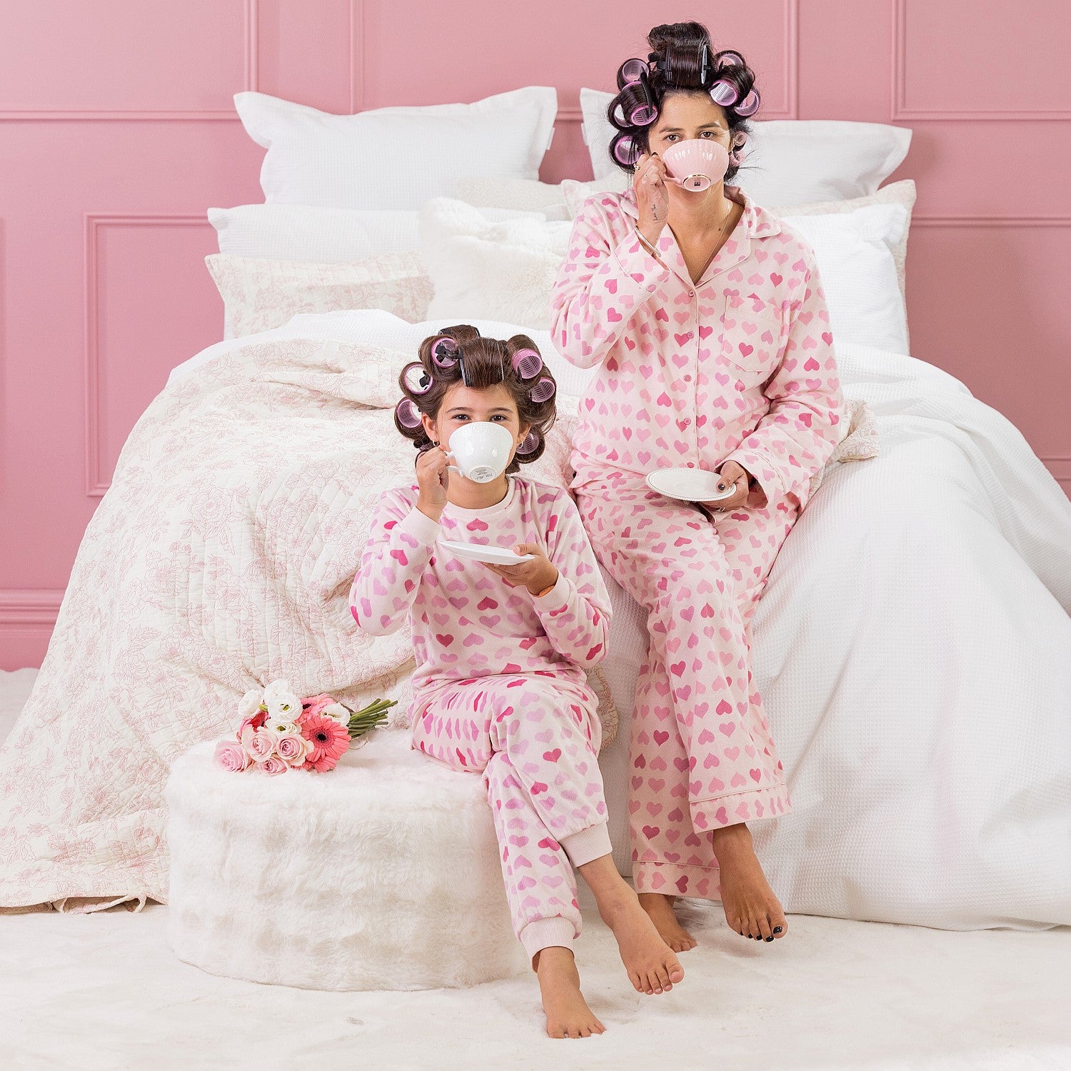 A kid, and mother wearing matching pink heart print pyjamas, sitting on a white bed with curlers in their hair.