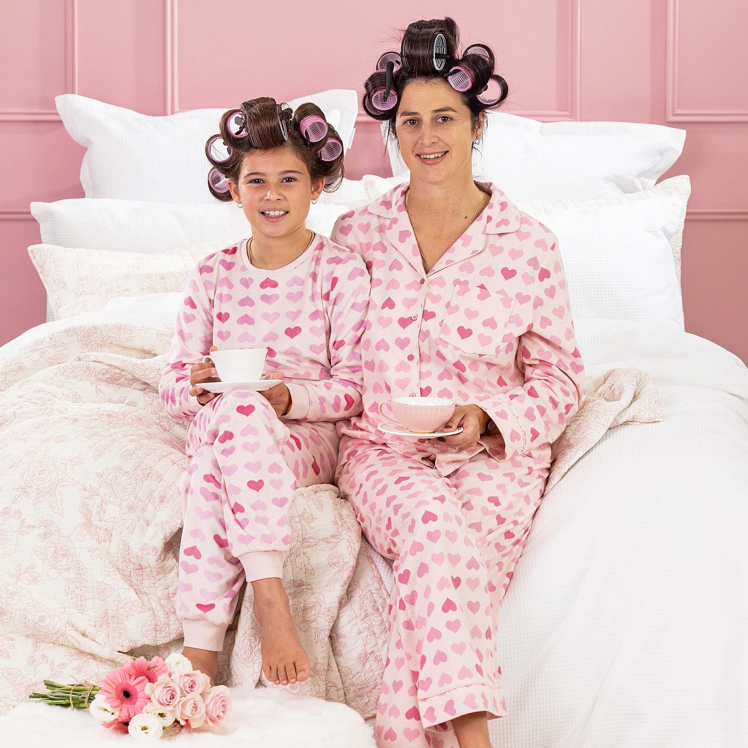A kid, and mother wearing matching pink heart print pyjamas, sitting on a white bed with curlers in their hair.