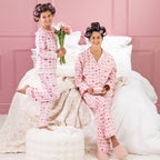 A kid, and mother wearing matching pink heart print pyjamas, sitting on a white bed with curlers in their hair.