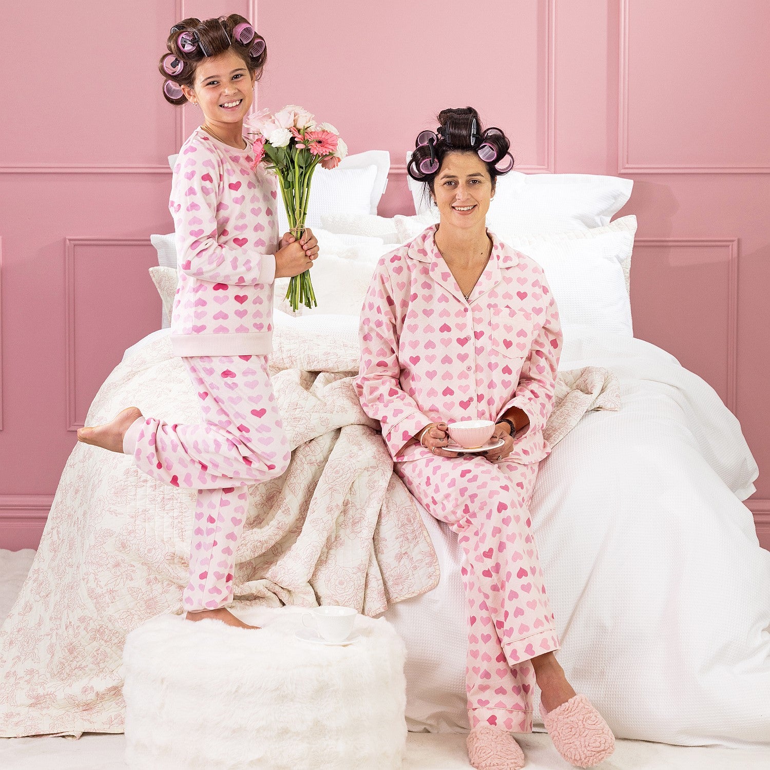 A kid, and mother wearing matching pink heart print pyjamas, sitting on a white bed with curlers in their hair.