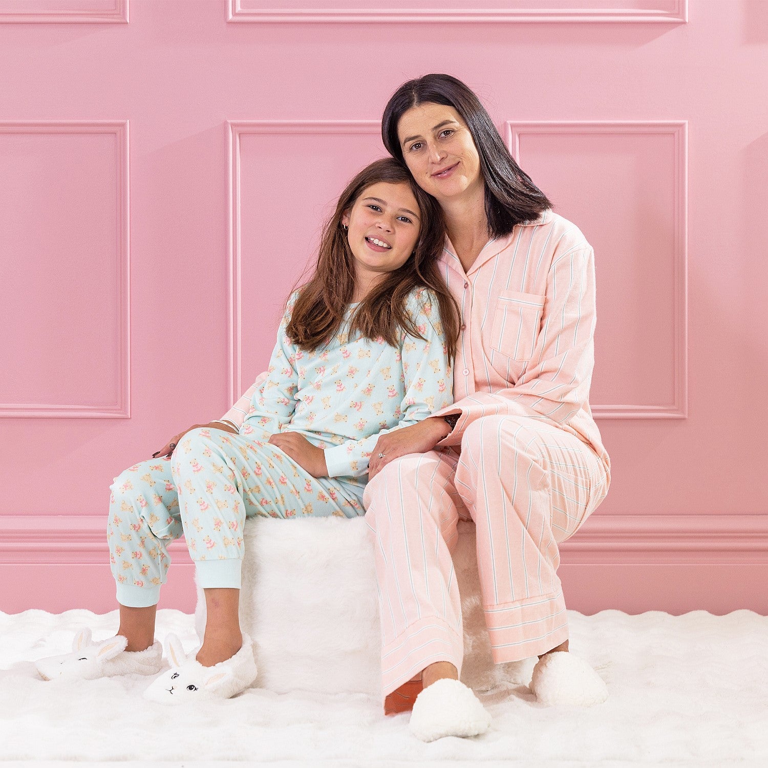children and adult wearing cosy pyjamas, sitting together on a white ottoman against a pink background.