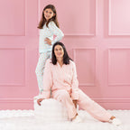 children and adult wearing cosy pyjamas, one standing and one sitting on a white ottoman against a pink background.