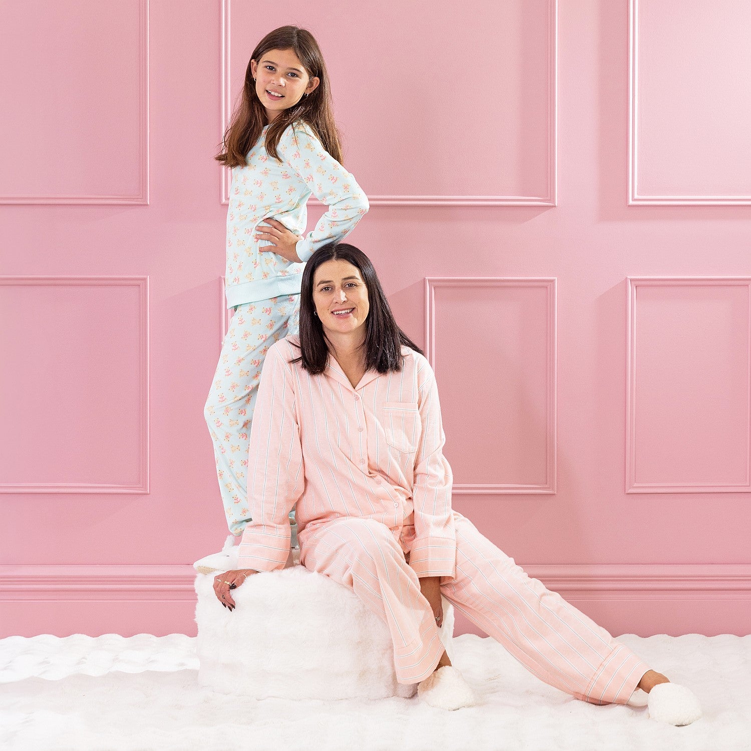 children and adult wearing cosy pyjamas, one standing and one sitting on a white ottoman against a pink background.