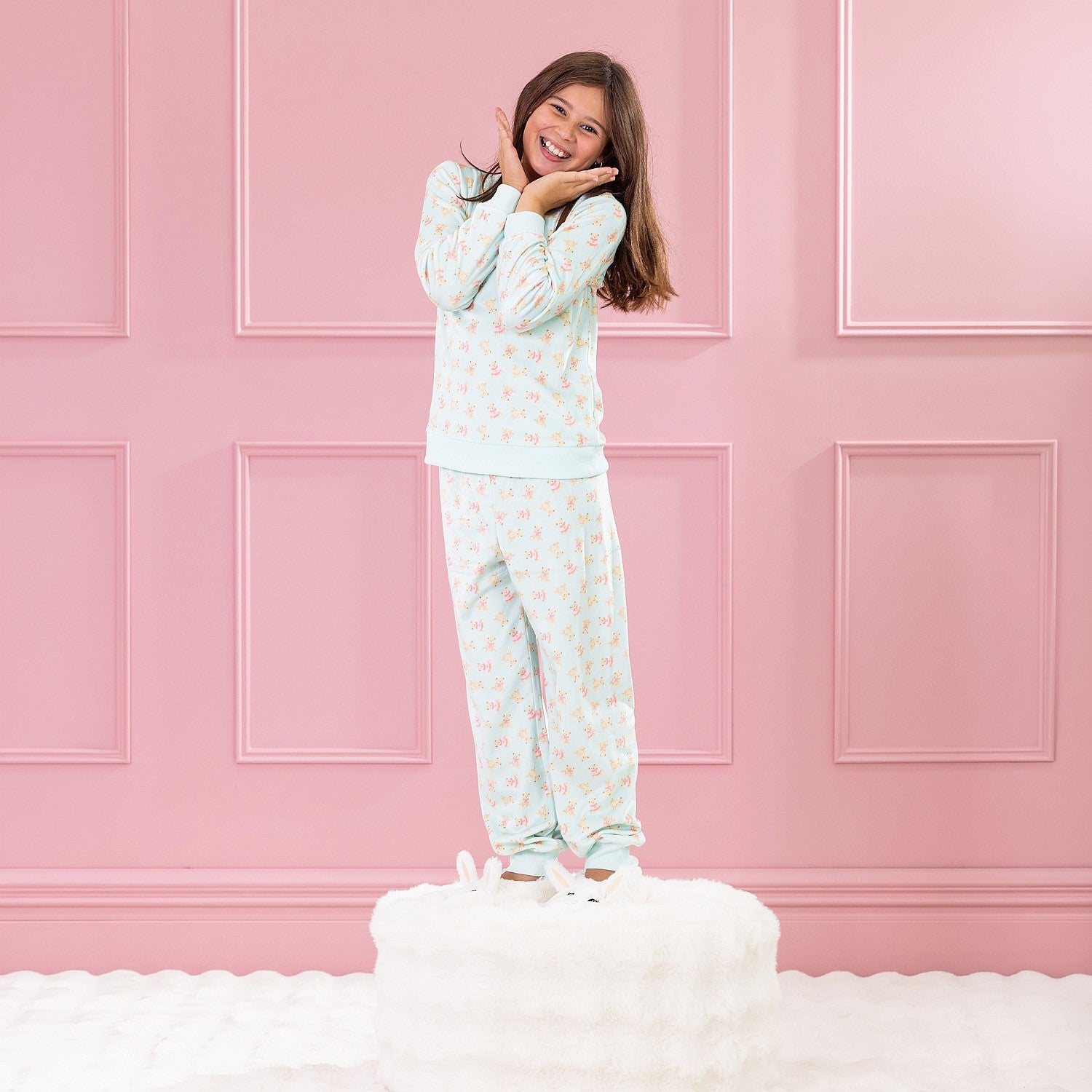 Child wearing light green teddy printed pyjamas standing on a white ottoman against a pink background.
