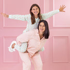 children and adult wearing cosy pyjamas, one standing and one on the others back  against a pink background.