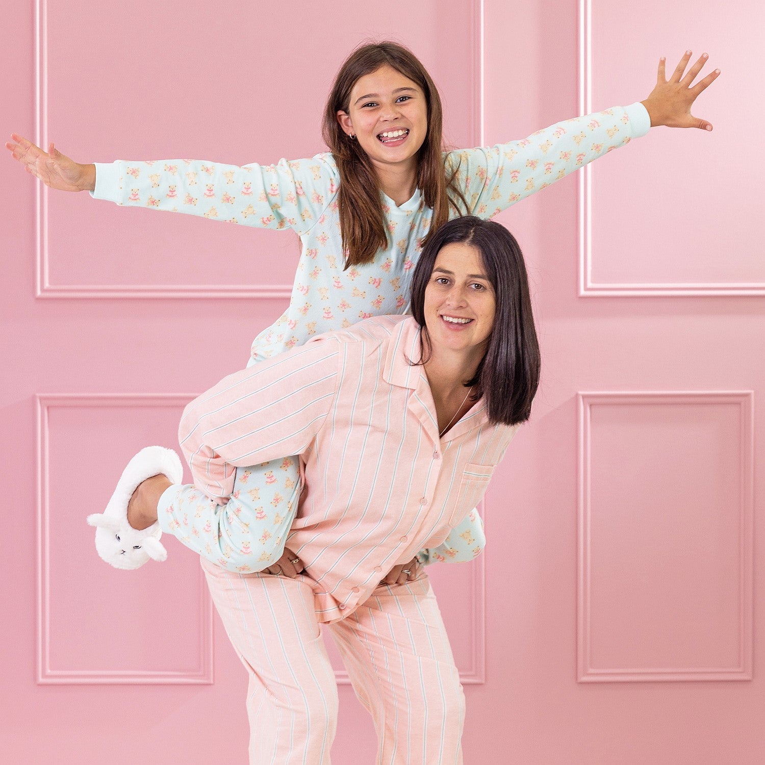 children and adult wearing cosy pyjamas, one standing and one on the others back  against a pink background.