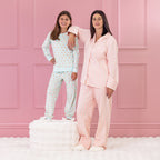 children and adult wearing cosy pyjamas, both standing, kid is on a white ottoman against a pink background.