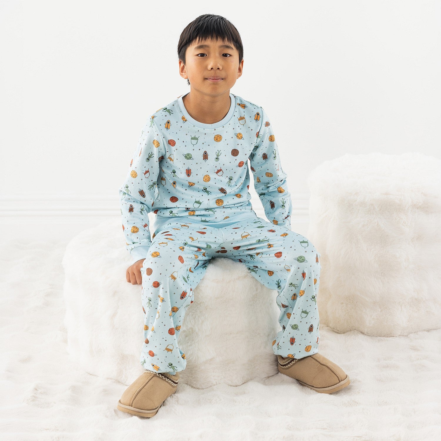 Child wearing light blue space printed pyjamas sitting on a stool on a white background.
