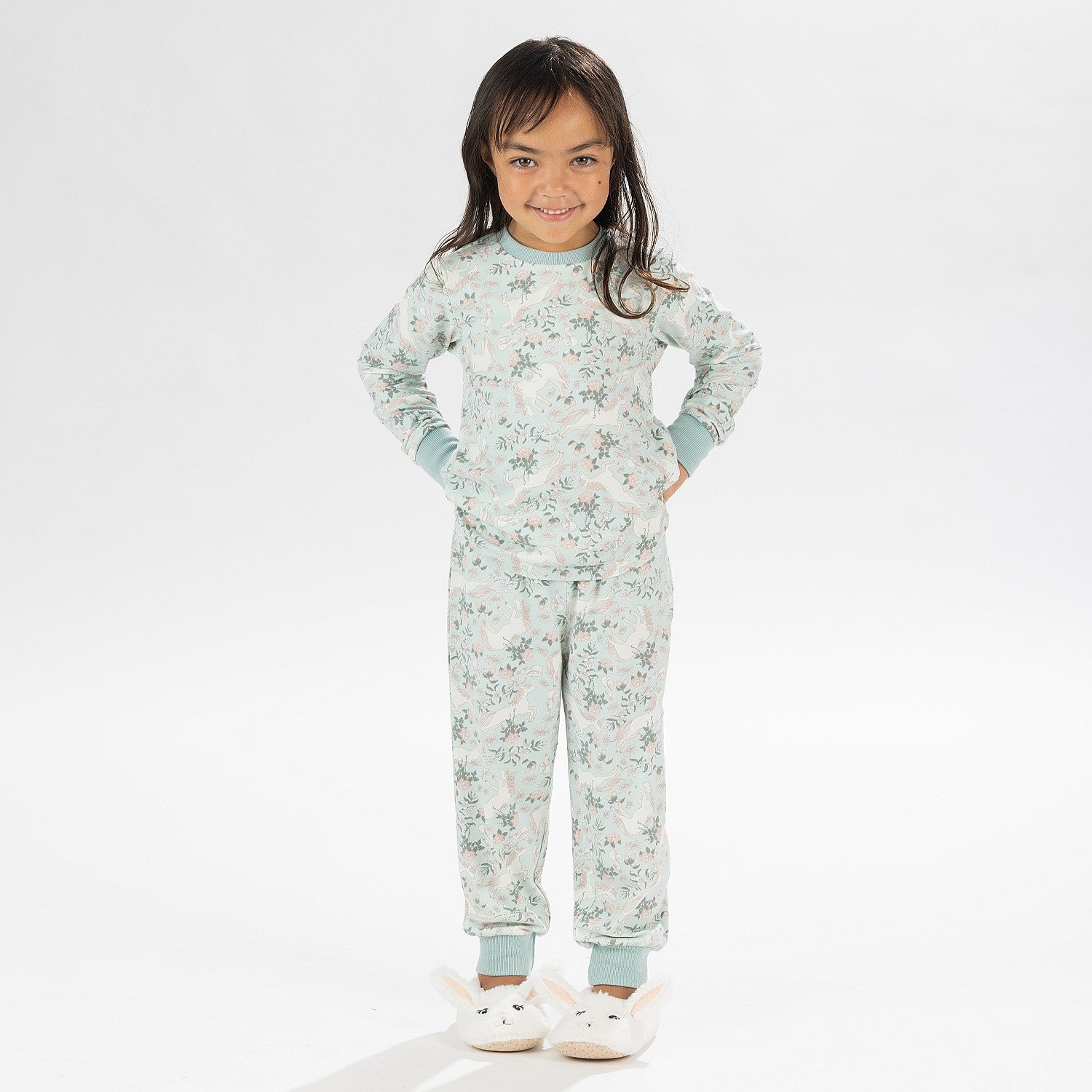 Child wearing a light green printed pyjama set with unicorns on a white background. Wearing bunny slippers.