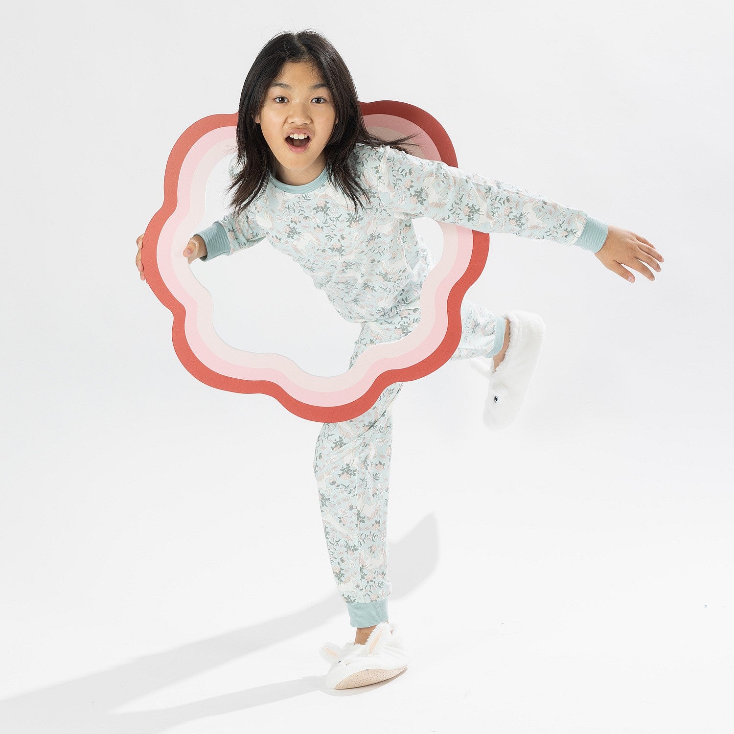 Child wearing a light green printed pyjama set with unicorns on a white background. Wearing bunny slippers.