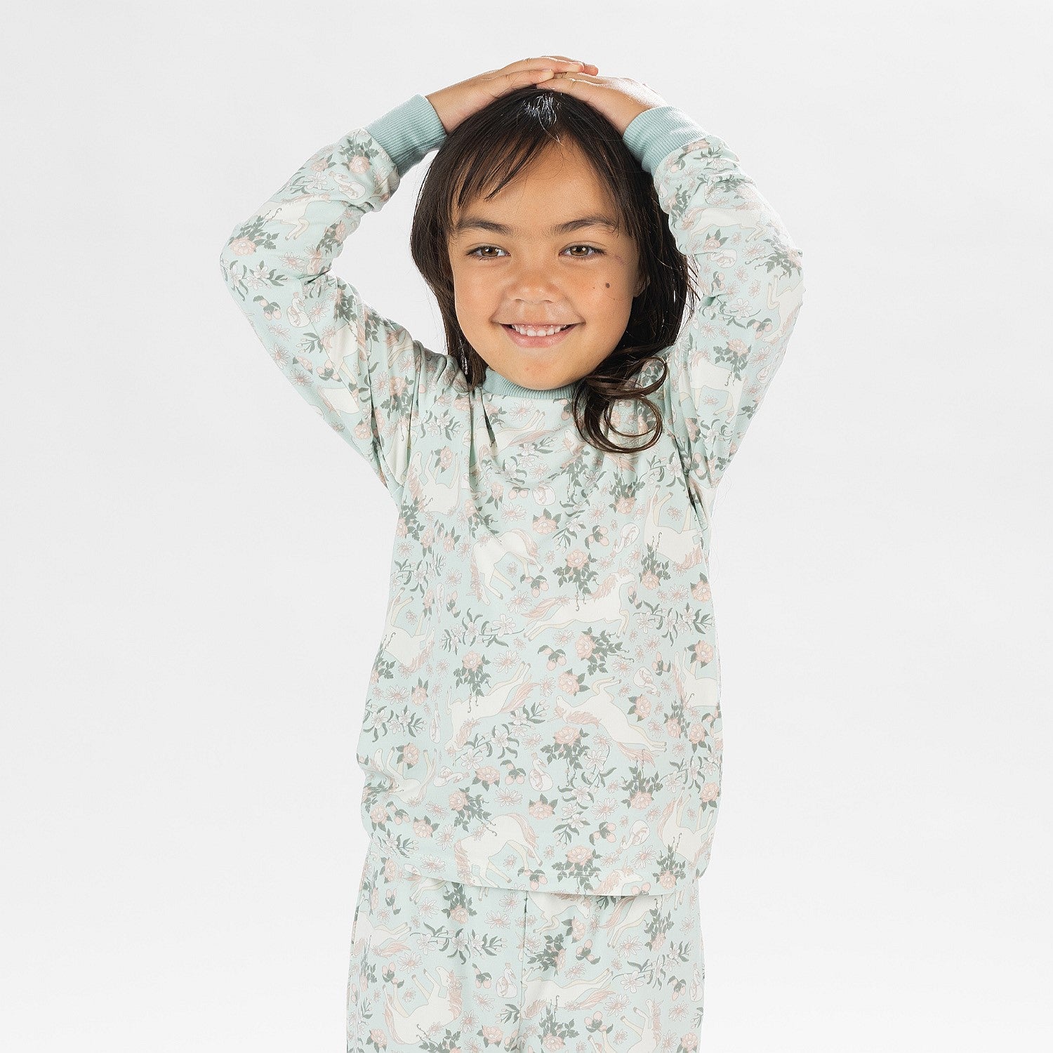 Child wearing a light green printed pyjama set with unicorns on a white background. Wearing bunny slippers.