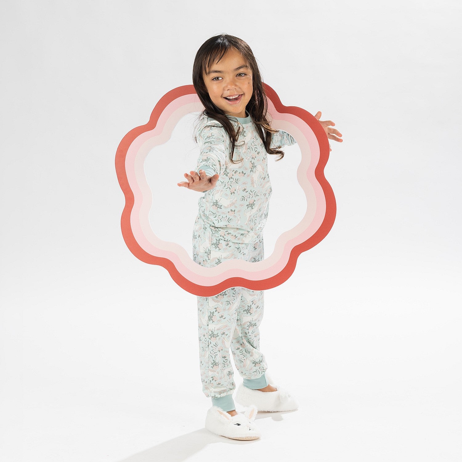 Child wearing a light green printed pyjama set with unicorns on a white background. Wearing bunny slippers.