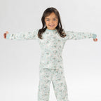 Child wearing a light green printed pyjama set with unicorns on a white background. Wearing bunny slippers.