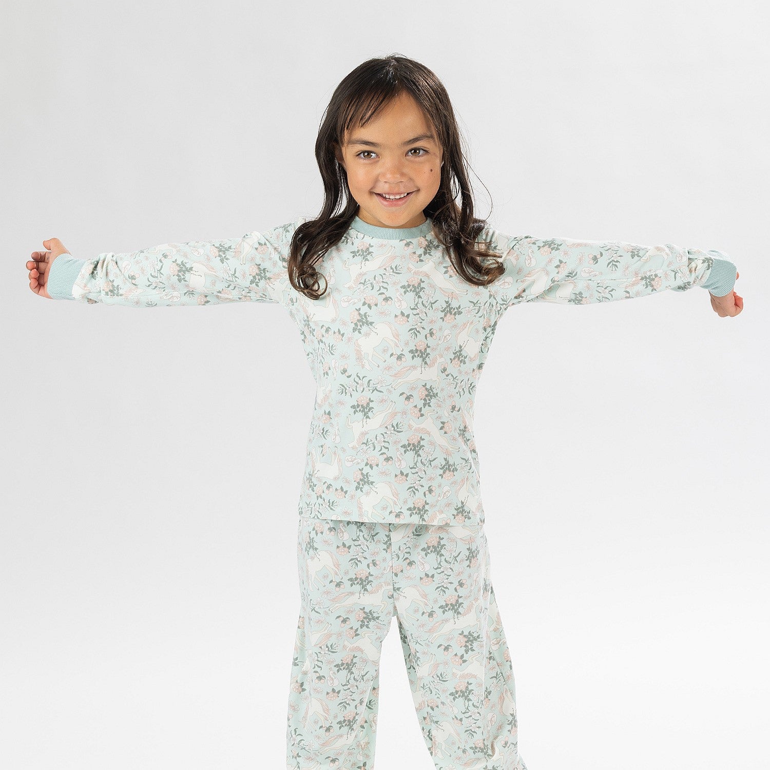 Child wearing a light green printed pyjama set with unicorns on a white background. Wearing bunny slippers.