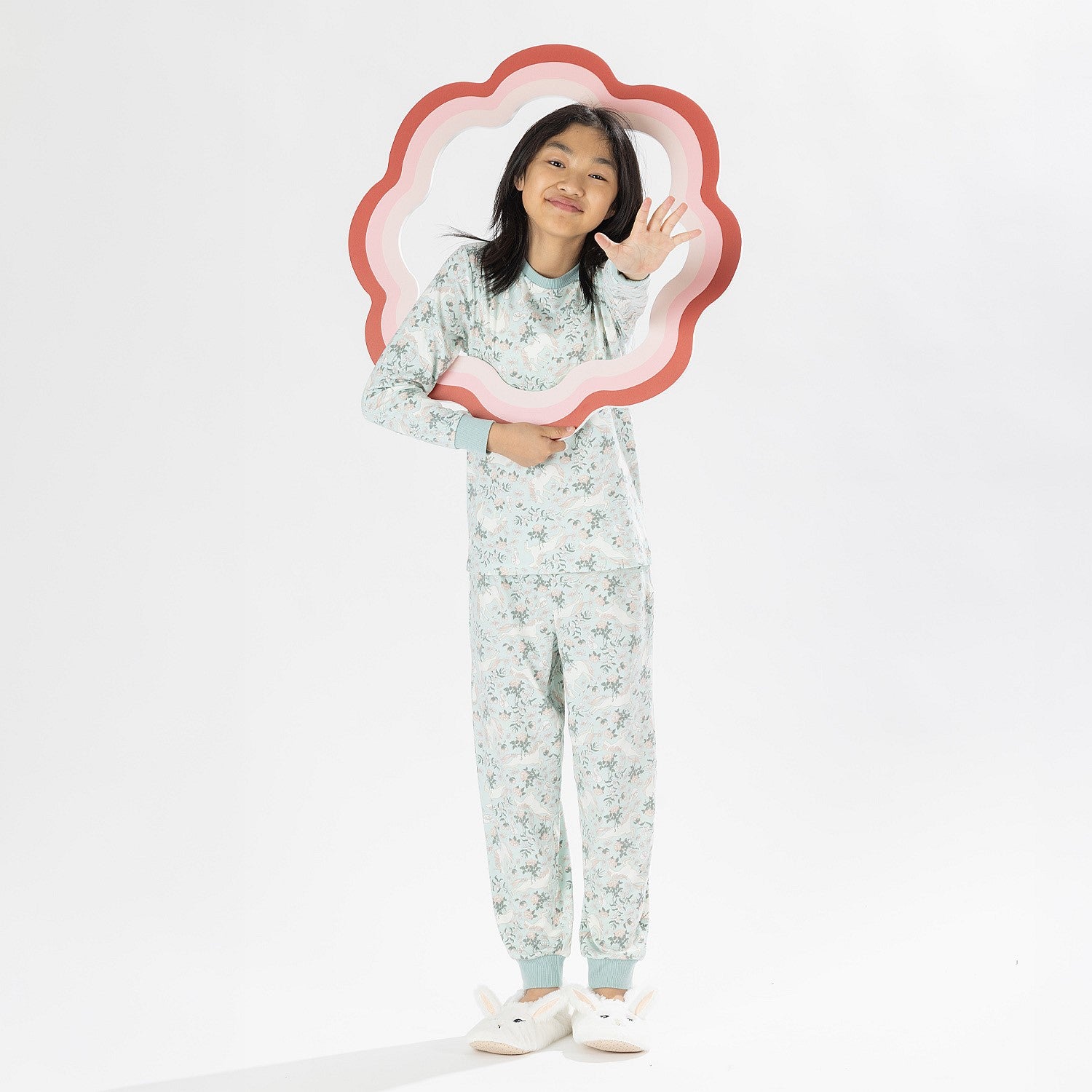 Child wearing a light green printed pyjama set with unicorns on a white background. Wearing bunny slippers.