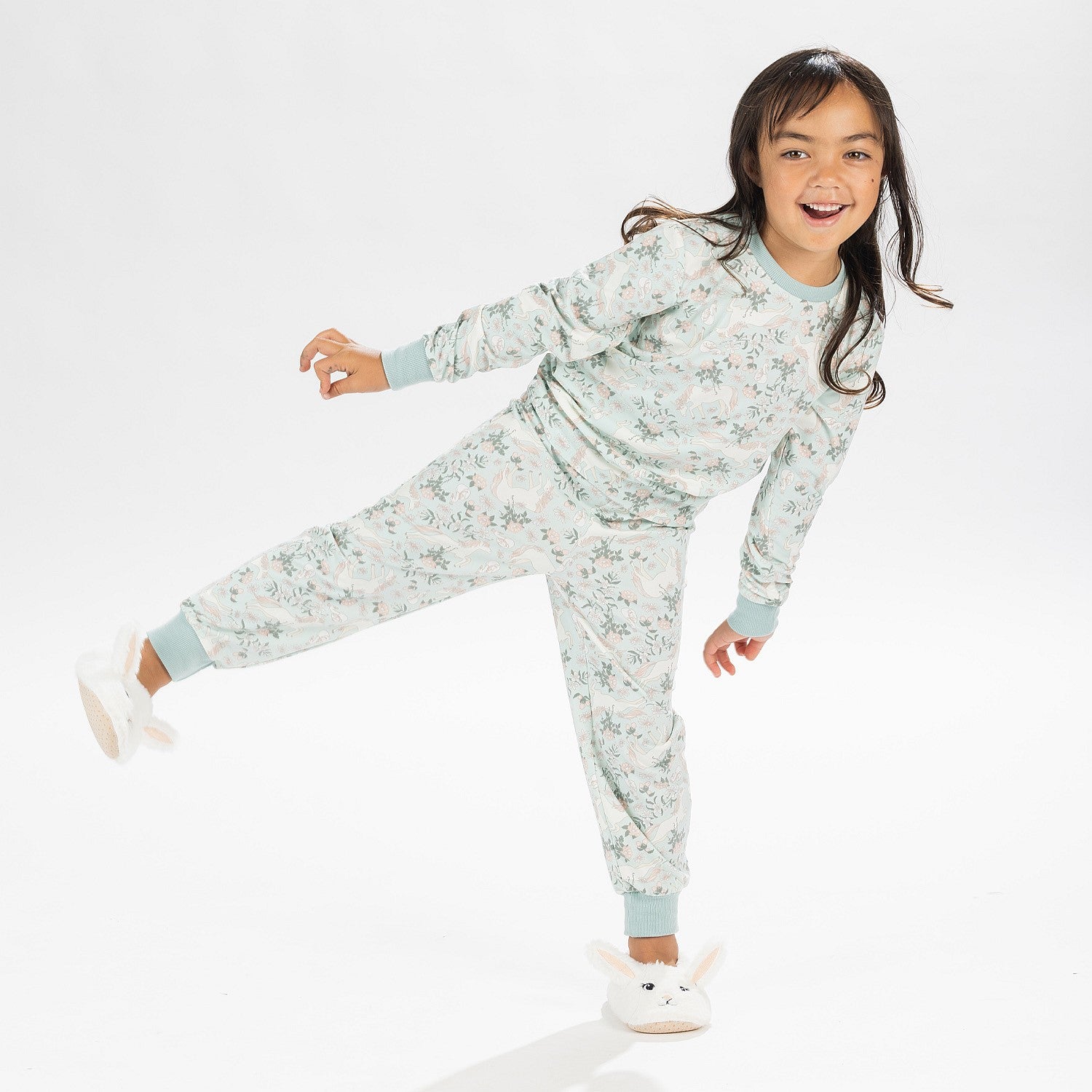 Child wearing a light green printed pyjama set with unicorns on a white background. Wearing bunny slippers.