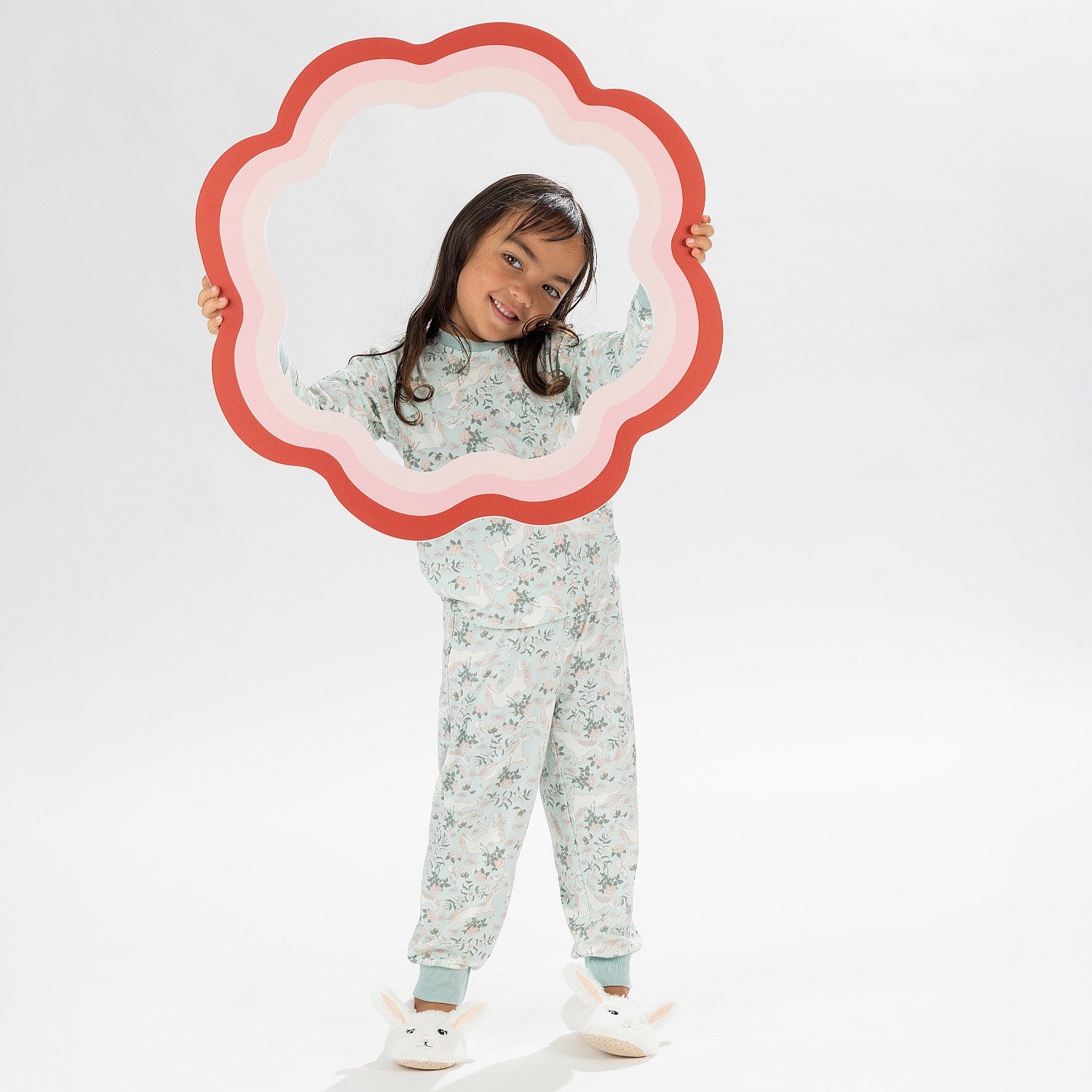 Child wearing a light green printed pyjama set with unicorns on a white background. Wearing bunny slippers.