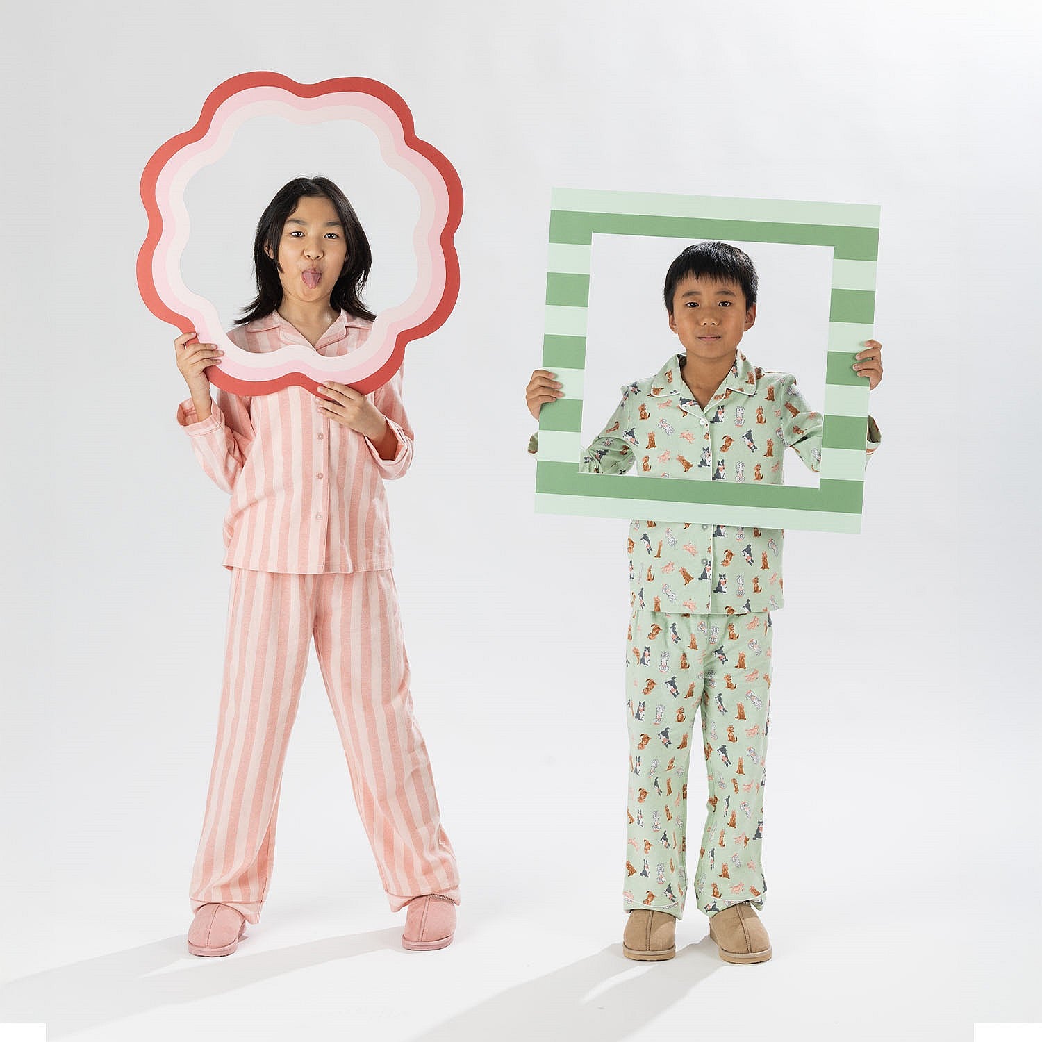Two kids wearing matching long sleeve pyjama sets, one in pink stripes and one in green print.
