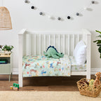 bb&b Kids Dino Beach Party cot styled with a mint green dinosaur-themed cot cover featuring playful dinosaurs, palm trees and beach motifs, with a plush dinosaur toy and white pillow.