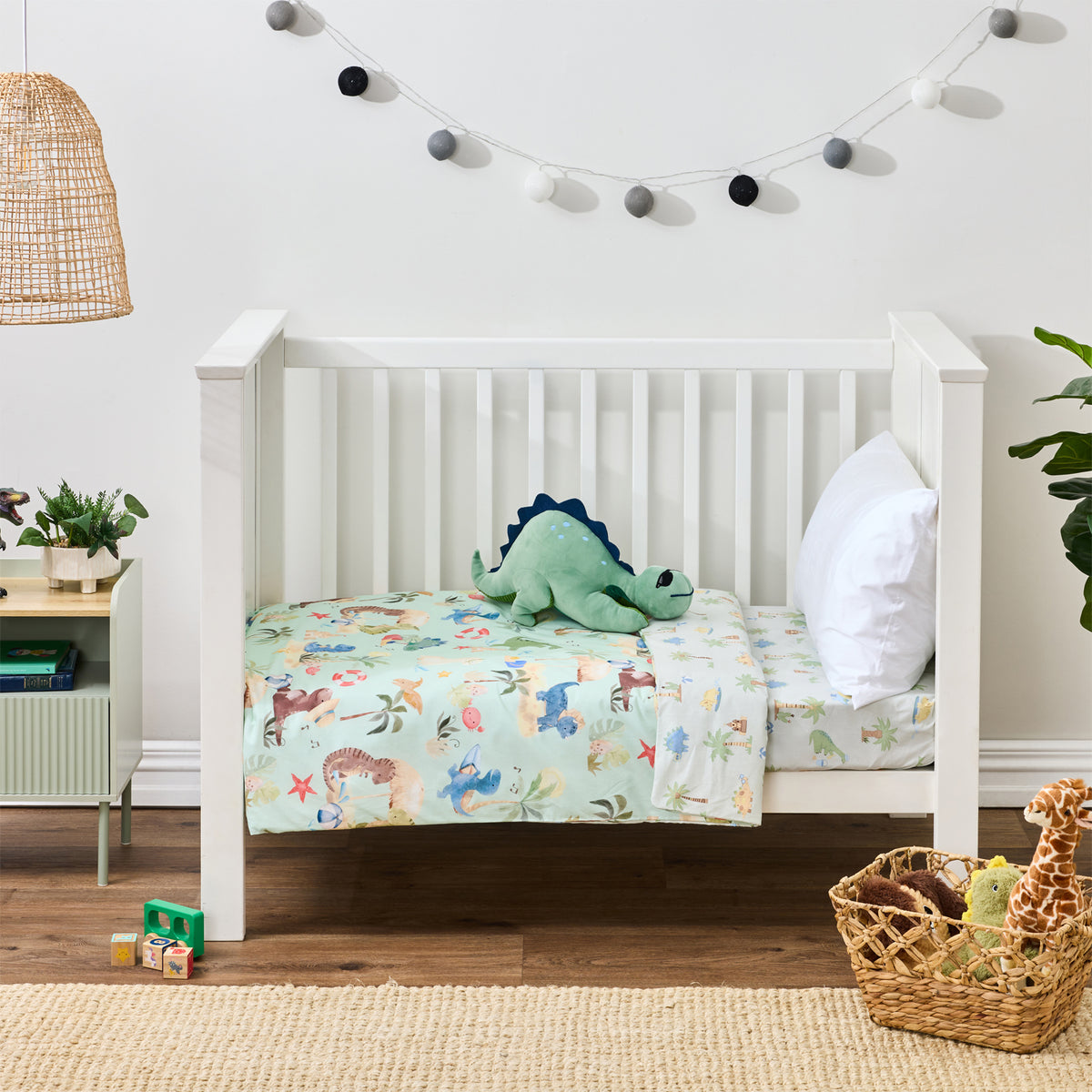 bb&b Kids Dino Beach Party cot styled with a mint green dinosaur-themed cot cover featuring playful dinosaurs, palm trees and beach motifs, with a plush dinosaur toy and white pillow.