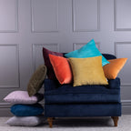 Assorted velvet cushions in bright colours styled on a navy chair against a grey panelled wall.