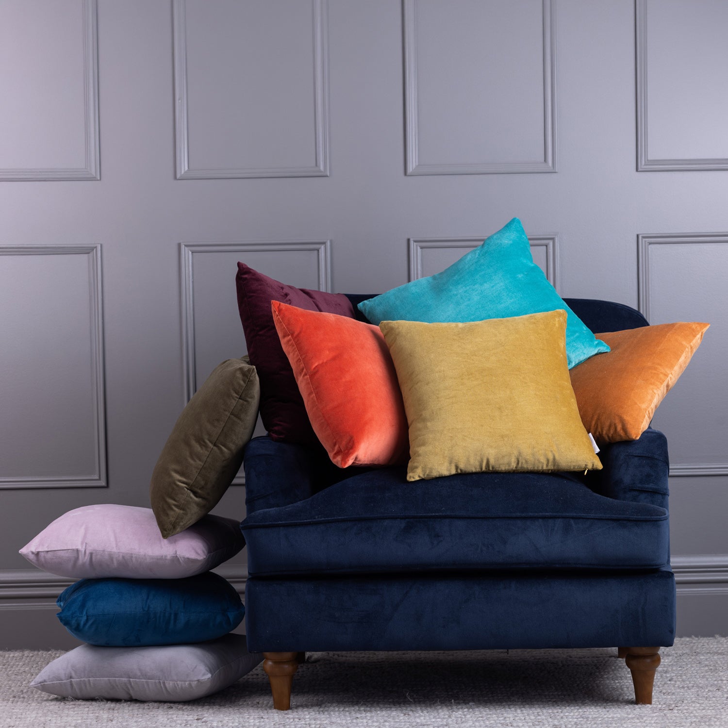 Assorted velvet cushions in bright colours styled on a navy chair against a grey panelled wall.
