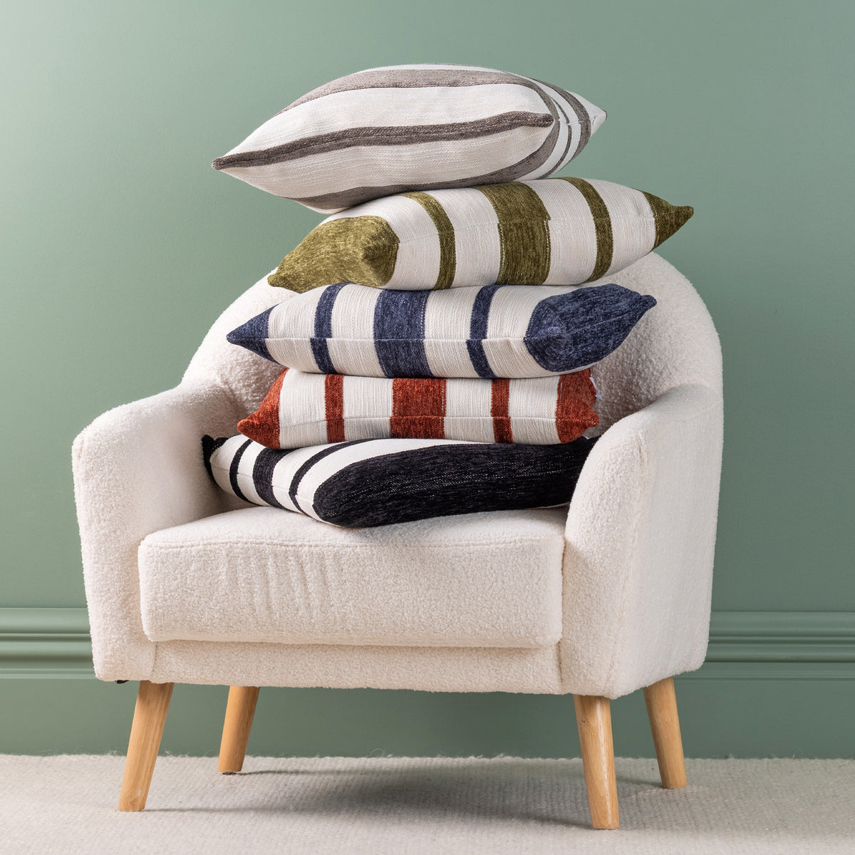 A stack of five decorative cushions in various striped and textured patterns sits on a cream boucle armchair with light wood legs, set against a sage green wall.