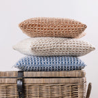 Three textured woven cushions in rust, natural, and blue tones are neatly stacked on top of a wicker storage basket, showcasing their rich tactile finish and coordinating colours.