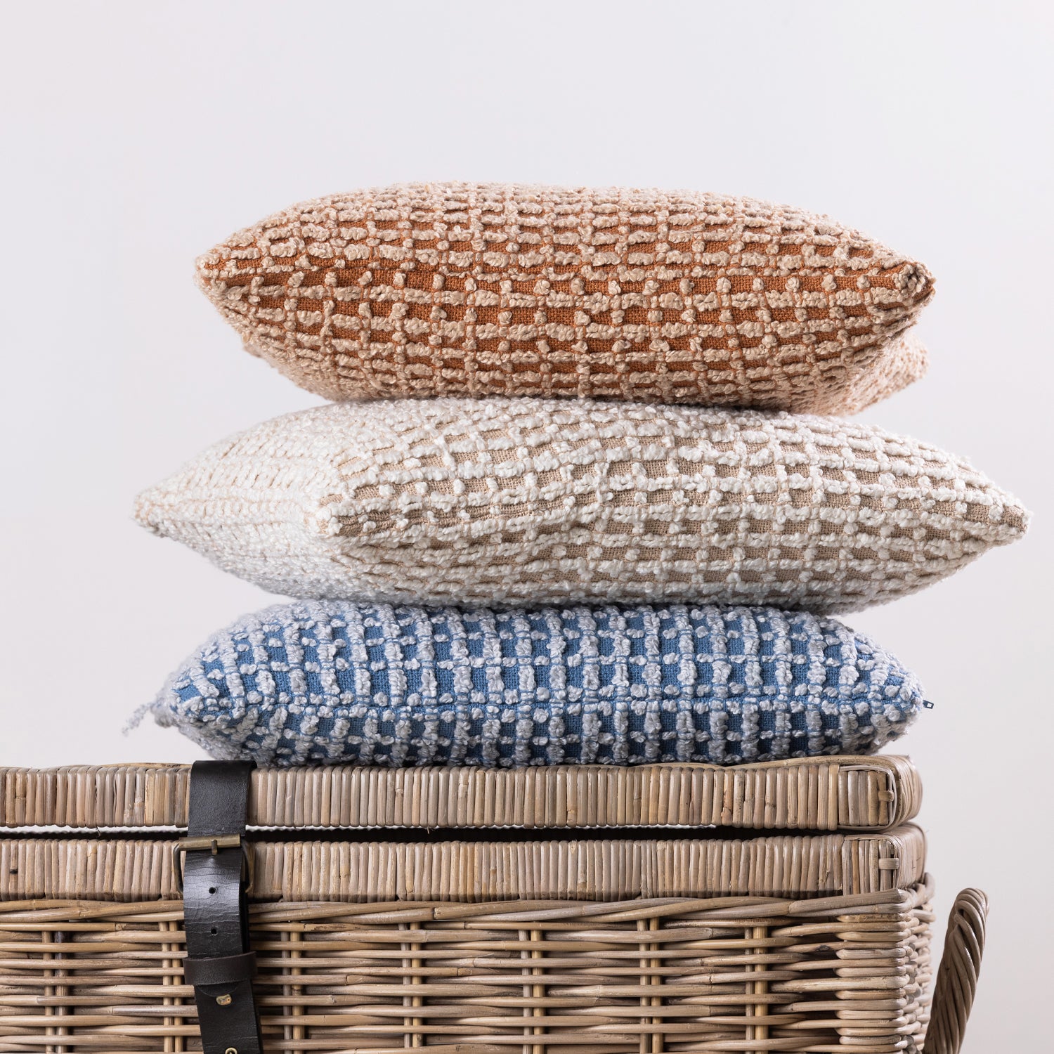 Three textured woven cushions in rust, natural, and blue tones are neatly stacked on top of a wicker storage basket, showcasing their rich tactile finish and coordinating colours.