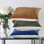 Three square quilted cushions in rust, olive green, and blue velvet, stacked on a black table next to a glass vase with white flowers.