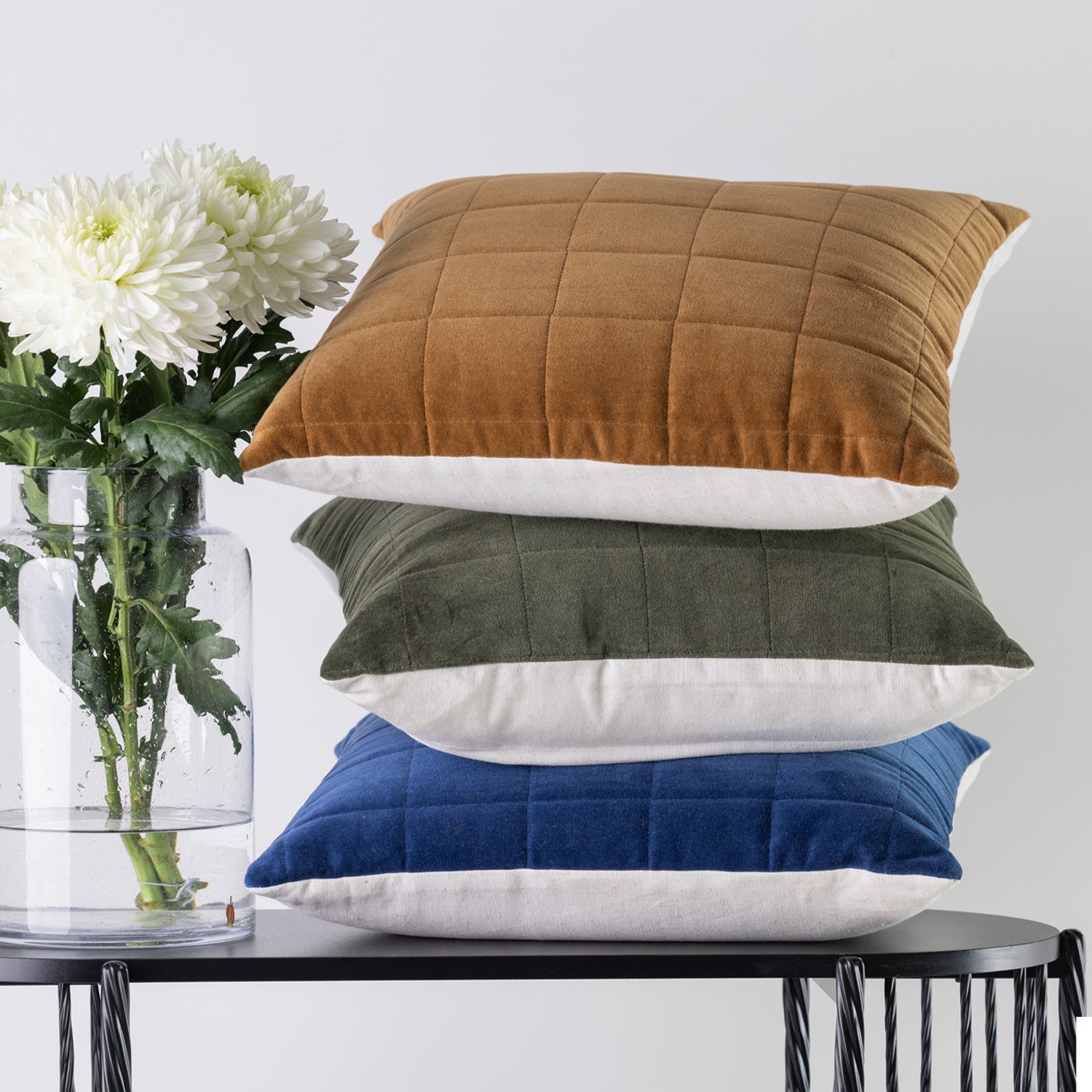 Three square quilted cushions in rust, olive green, and blue velvet, stacked on a black table next to a glass vase with white flowers.