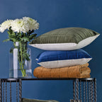 A black metal table styled with rust, blue, olive green cushions and throws. Staged with a clear glass vase of white flowers, set against a dark blue wall.