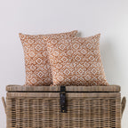 Two square throw pillows with a rust-orange and cream geometric pattern, displayed on top of a rustic wicker storage chest against a plain white wall.