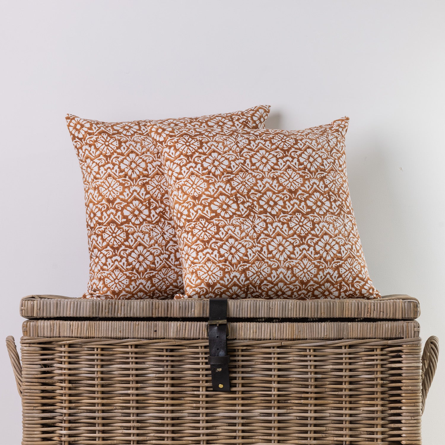 Two square throw pillows with a rust-orange and cream geometric pattern, displayed on top of a rustic wicker storage chest against a plain white wall.