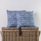 Two square throw pillows with a blue and white floral pattern, displayed on top of a large woven wicker storage trunk against a plain light gray wall.
