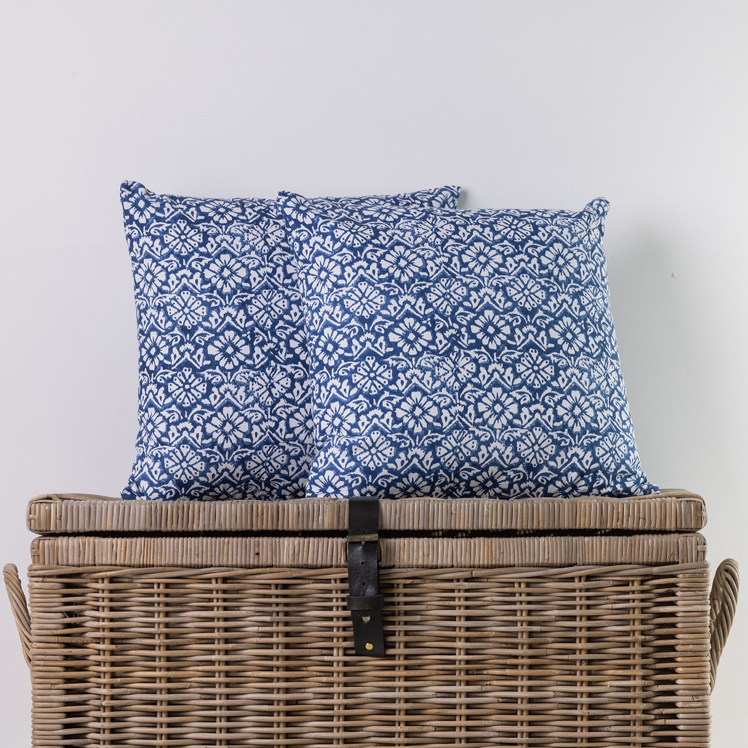 Two square throw pillows with a blue and white floral pattern, displayed on top of a large woven wicker storage trunk against a plain light gray wall.