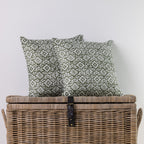 Two square throw pillows with an olive green and white geometric pattern, displayed on top of a brown woven wicker storage trunk against a plain light gray wall.