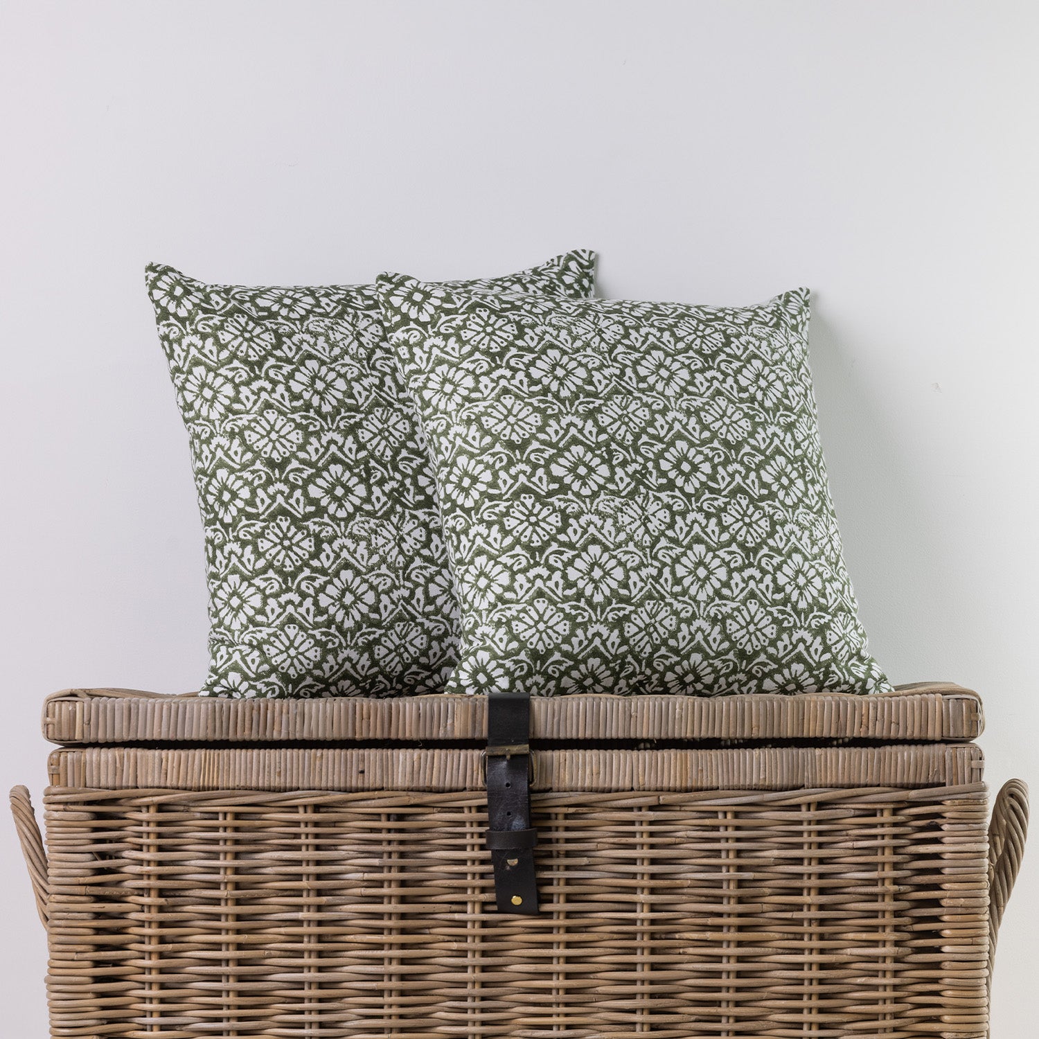 Two square throw pillows with an olive green and white geometric pattern, displayed on top of a brown woven wicker storage trunk against a plain light gray wall.