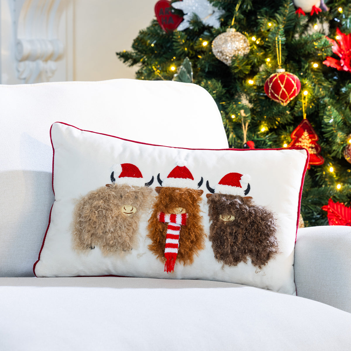 Highland cows Christmas cushion displayed on a white sofa beside a Christmas tree with festive ornaments and warm fairy lights.