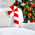 A red and white textured candy cane cushion displayed on a white armchair beside a decorated Christmas tree.