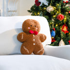 A brown textured gingerbread man cushion with a red bow tie, embroidered face and white icing details, displayed on a white armchair beside a Christmas tree.