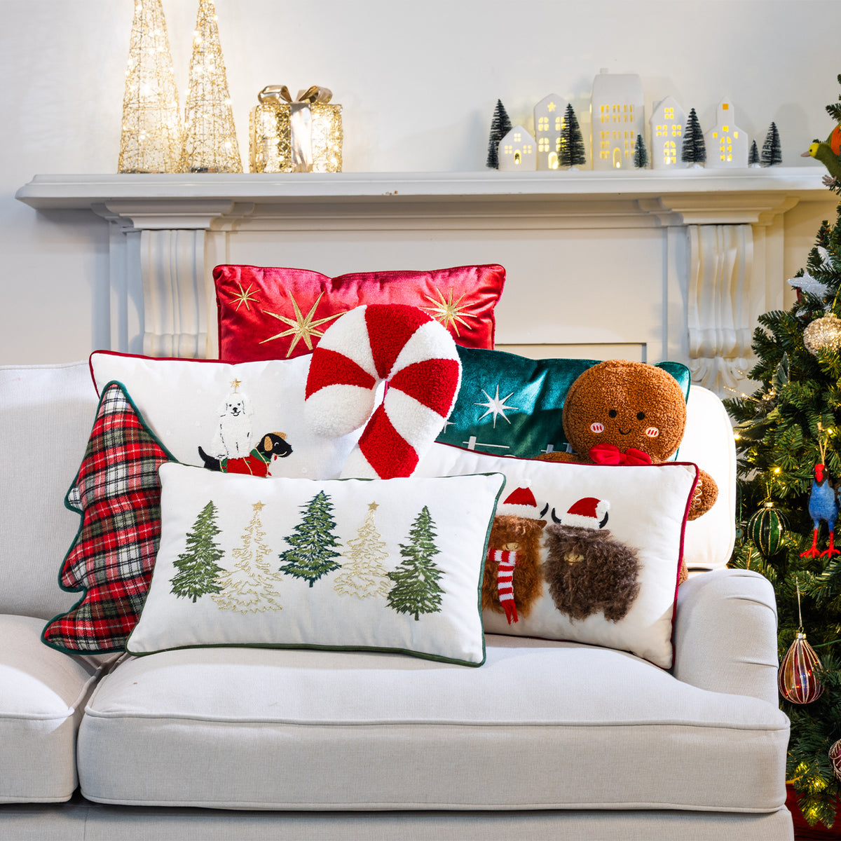 A collection of festive Christmas cushions arranged on a white sofa beside a decorated Christmas tree and fireplace.