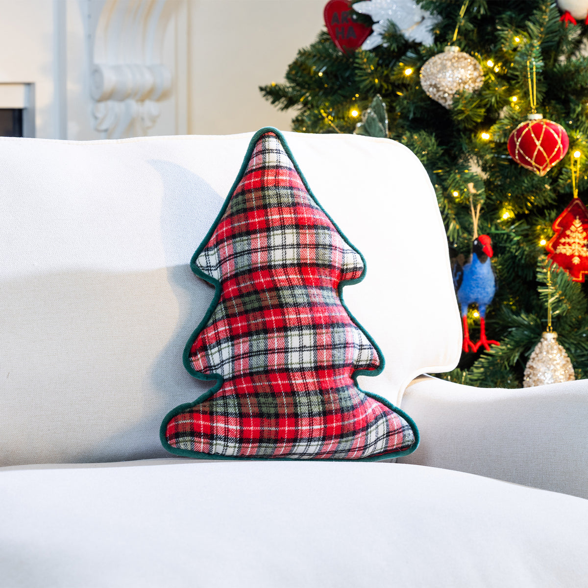 Red, green and white check Christmas tree-shaped cushion with dark green piping, displayed on a white sofa beside a decorated Christmas tree.